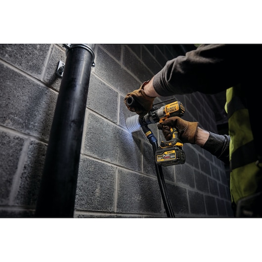 A person wearing work gloves and a safety vest is using a DEWALT cordless drill with a DCB606 FLEXVOLT battery to drill into a concrete block wall, with a dust extraction attachment connected.