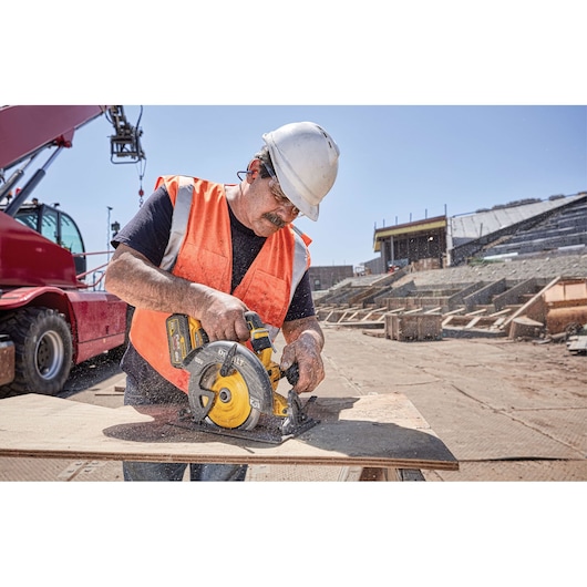 A construction worker wearing a white hard hat and orange safety vest uses a yellow and black DEWALT circular saw to cut a wooden board at an outdoor construction site. A red heavy machine and building structures are visible in the background.
