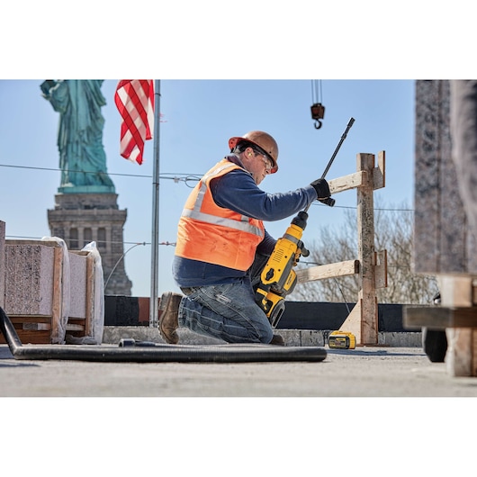 A construction worker wearing a hard hat and orange safety vest uses a yellow DEWALT power tool outdoors, with the Statue of Liberty and an American flag visible in the background.