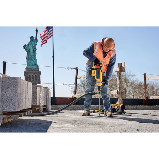 A construction worker wearing a hard hat and orange safety vest is using a yellow DEWALT power tool to drill into a concrete surface at a job site. Granite blocks and construction materials are visible nearby, with the Statue of Liberty and an American flag in the background.