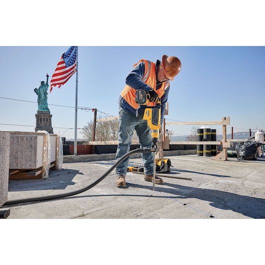 A construction worker wearing a hard hat and orange safety vest uses a yellow DEWALT power tool to drill into concrete at a work site. The Statue of Liberty and an American flag are visible in the background.