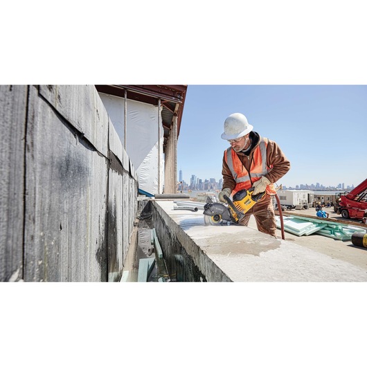 A construction worker wearing a white hard hat and orange safety vest operates a DEWALT power tool to cut concrete on a building site. The city's skyline is visible in the background.