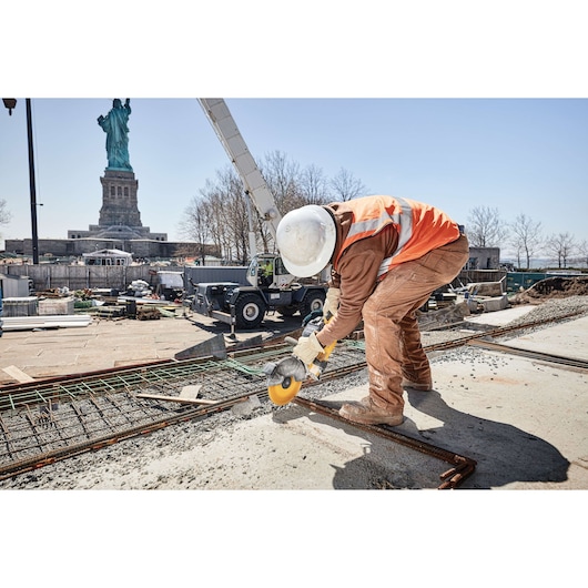 A construction worker wearing a hard hat, gloves, and an orange safety vest is using a DEWALT power tool to cut metal rebar at a construction site. The Statue of Liberty is visible in the background.