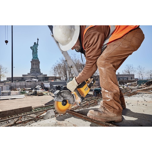 A construction worker uses a DEWALT cordless circular saw to cut through metal rebar at a construction site. The worker is wearing a white hard hat, orange safety vest, gloves, and brown work clothes. The Statue of Liberty is visible in the background on a clear day.