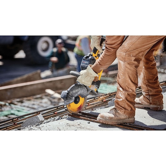 A worker wearing protective gloves and brown work clothes is using a DEWALT power saw to cut metal rebar on a construction site.