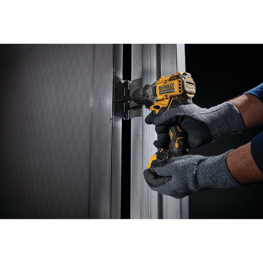 A person wearing gray gloves uses a yellow and black DEWALT cordless drill to secure a hinge on a metal door frame.