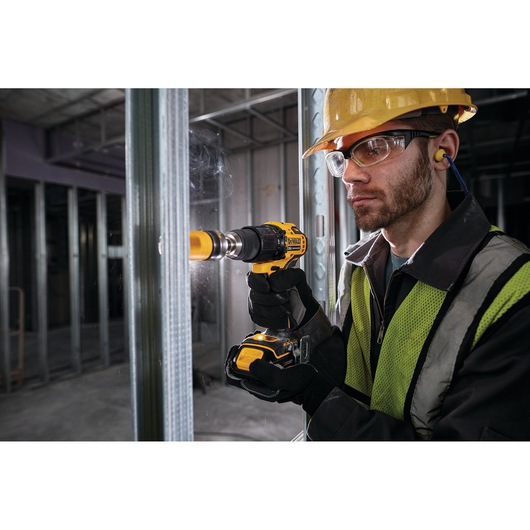 A person wearing safety gear and a yellow vest is using a DEWALT cordless drill (model DCD709B) with a hole saw bit to drill into a metal stud in a construction environment.