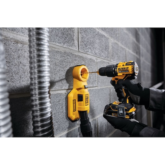 A DEWALT cordless drill is being used to drill into a concrete block wall. The drill is held by a person wearing black gloves and a DEWALT dust extraction attachment is fixed to the wall beneath the drill bit.