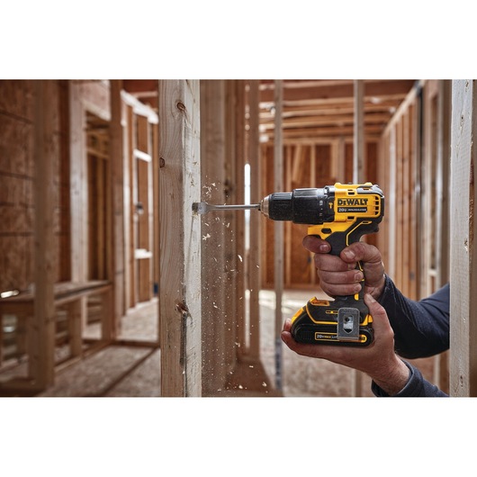 A DEWALT cordless power drill (model DCD709B) being used to drill into a wooden stud inside a building under construction. Wood shavings are visible as the drill bit penetrates the wood.