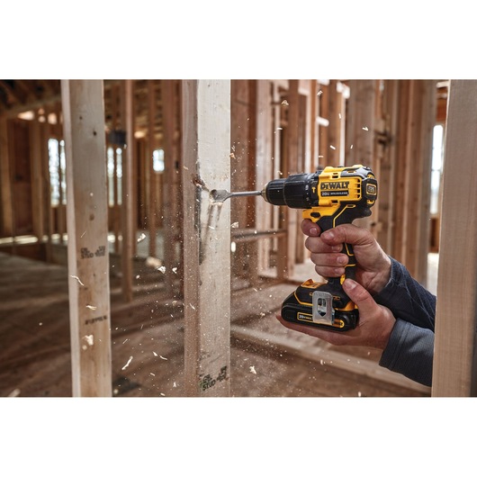 A DEWALT cordless drill (model DCD709B) being used to drill a hole in a wooden beam inside a construction site. Wood shavings are flying as the drill operates.