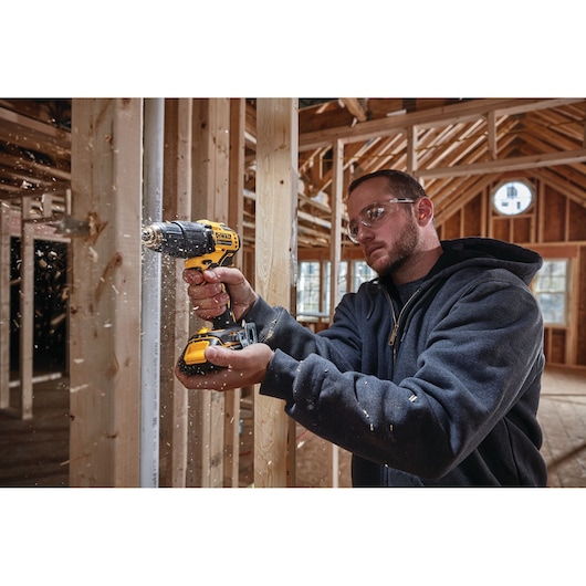 A person using a DEWALT cordless drill (model DCD709B) to drill into a wooden stud inside a building under construction. Wood shavings are flying as the drill is in use. The person is wearing a dark hoodie and their face is blurred.