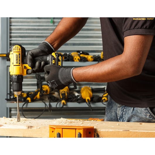 A person wearing black gloves is using a yellow DEWALT cordless drill to bore a hole into a wooden plank. In the background, several other yellow power tools are organized on a shelf.