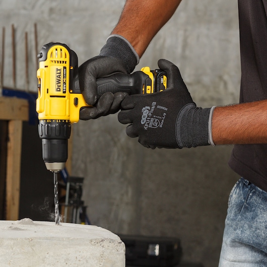 A person wearing black gloves is using a yellow DEWALT cordless drill to drill into a concrete surface.