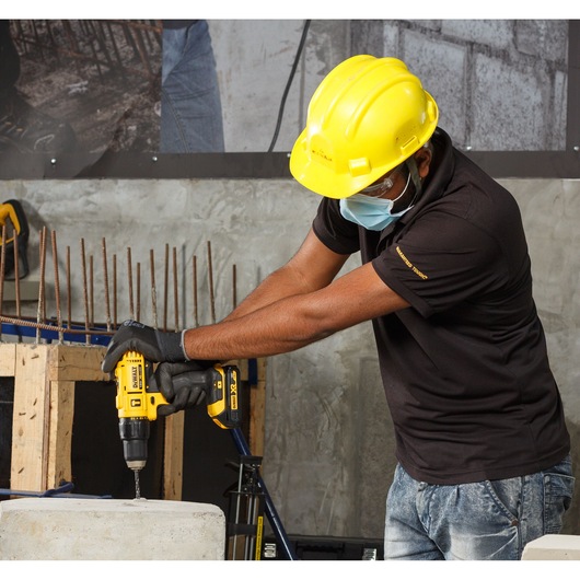 A person wearing a yellow safety helmet and black gloves operates a DEWALT DCD776S2 cordless drill, drilling into a concrete block at a construction site. The person's face is blurred for privacy.