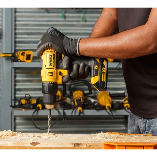 A person wearing gloves is using a yellow DEWALT DCD776S2 cordless drill to drill into a wooden surface, with wood shavings scattered around. Several yellow DEWALT power tools are visible in the background on a shelf.