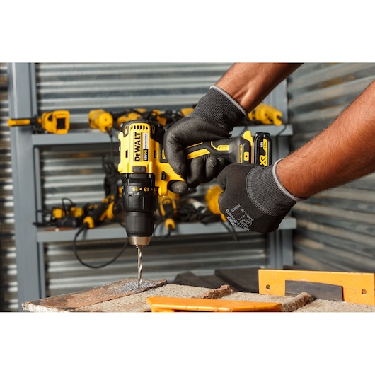 A person wearing work gloves is using a yellow and black DEWALT cordless drill to drill into a piece of metal in a workshop. In the background, several other DEWALT power tools are visible on shelves.