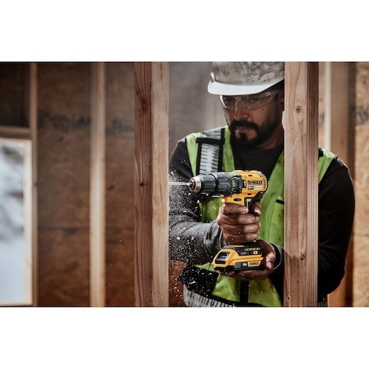 A person wearing a safety vest and helmet is using a DEWALT cordless power drill to make a hole in a wooden beam at a construction site. Wood shavings are visible around the drill bit.