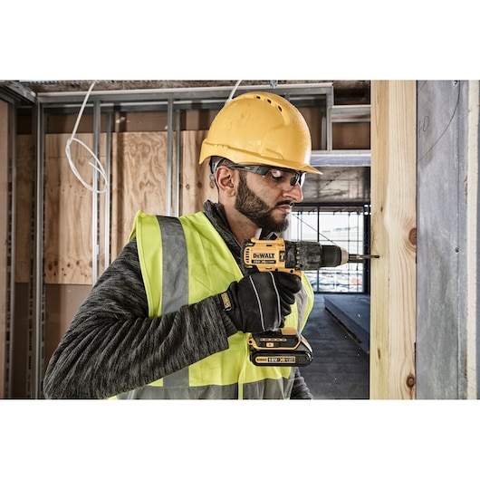 A person wearing a yellow safety helmet and high-visibility vest is using a DEWALT DCD778S2T cordless drill to drill into a wooden plank at a construction site. The person's face is obscured for privacy.