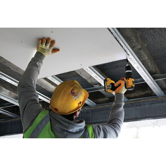 A construction worker wearing a yellow hard hat and safety vest uses a DEWALT cordless drill to attach a panel to the ceiling framework.