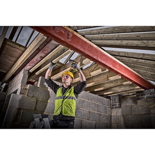 A construction worker wearing a yellow safety helmet and a green reflective vest is using a DEWALT cordless drill to fasten wooden beams to a red steel support inside a building under construction. The walls are made of concrete blocks and wooden rafters are visible above.