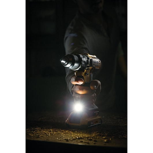 A person holding a DEWALT cordless power drill with a built-in LED light, illuminating a dark workspace. Wood shavings are visible on the surface below.