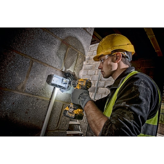 A construction worker wearing a yellow safety helmet and high-visibility vest is using a DEWALT cordless drill (DCD796N) to install or fix an electrical box onto a concrete wall. The worker is wearing protective gloves and the drill's LED light is illuminating the work area.