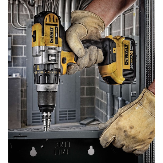 A close-up view of a person wearing work gloves using a yellow and black DEWALT cordless drill to create a hole in a metal surface, with an industrial setting in the background.