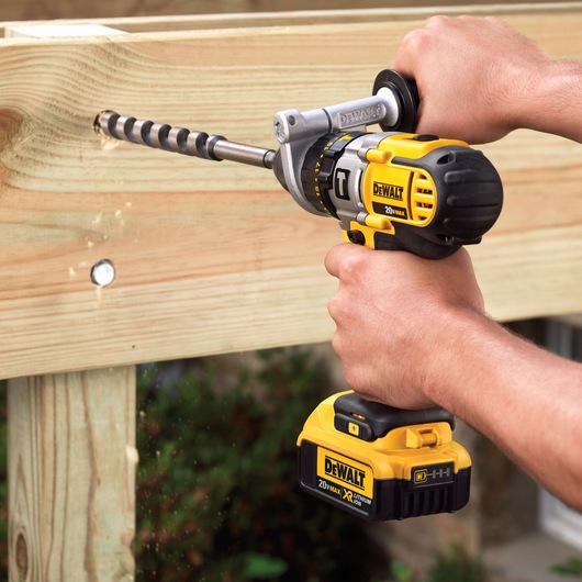 A person using a DEWALT 20V Max cordless power drill with a large drill bit to drill into a wooden plank.