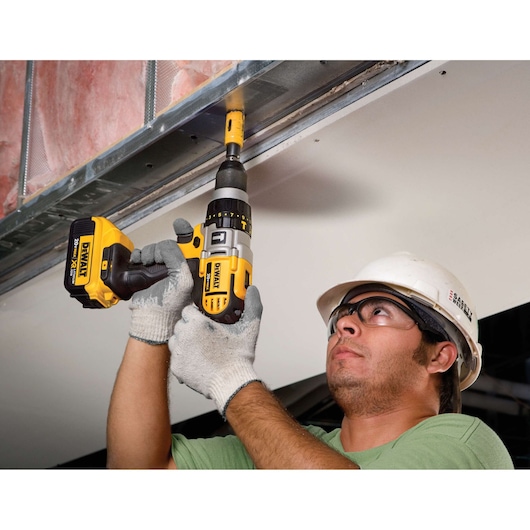 A worker wearing gloves and a hard hat is using a DEWALT DCD985M2 cordless drill to drive a screw into a metal frame above his head. The drill is yellow and black, and the worker is installing drywall or sheet material in a construction setting.