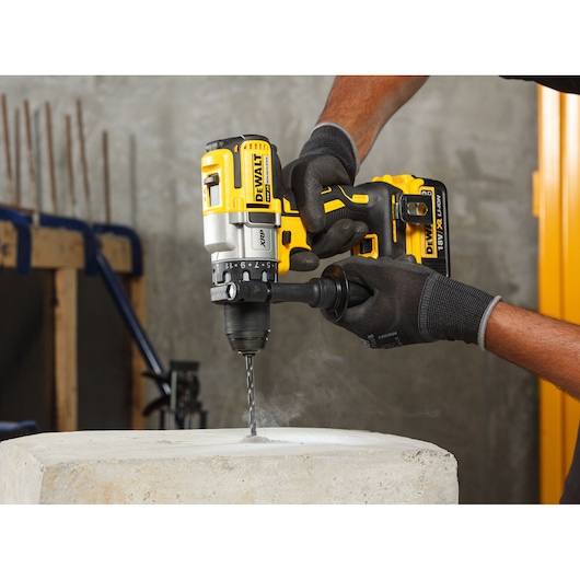 A person wearing black gloves is using a yellow DEWALT DCD991NT cordless drill to drill into a concrete block in a workshop setting.