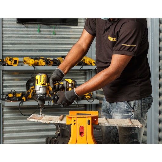 A person wearing gloves and a black DEWALT-branded polo shirt uses a DEWALT DCD991NT cordless drill to drill into a piece of material, with several other DEWALT power tools visible on shelves in the background.