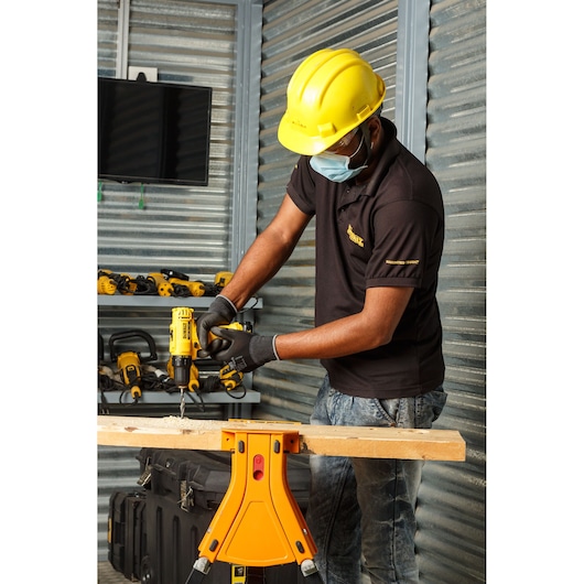 A person wearing a yellow hard hat and black shirt is using a DEWALT cordless drill to drill into a piece of wood in a workshop. The workshop has metallic walls, shelves with various DEWALT power tools, and a workbench.