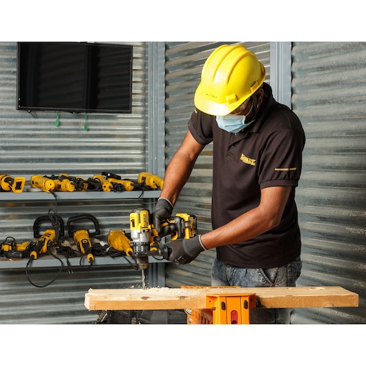 A worker wearing a yellow hard hat, protective gloves, and a face mask uses a DEWALT cordless drill to bore into a piece of wood in a workshop. Other DEWALT power tools are visible on shelves in the background.