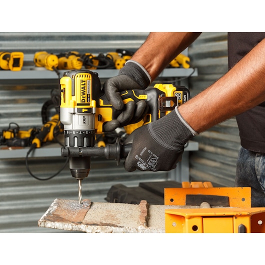 A person wearing protective gloves is using a yellow DEWALT cordless drill to drill into a metal surface. The drill is held securely, and there are other power tools visible on shelves in the background.