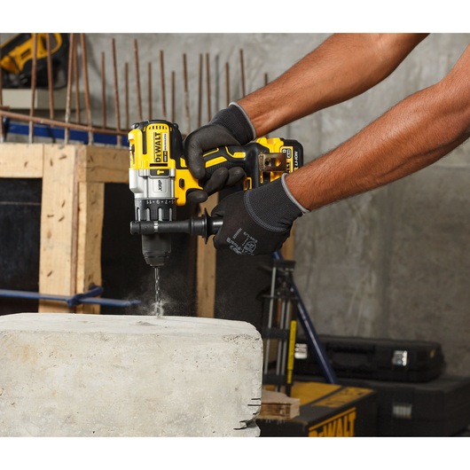 A person wearing black work gloves uses a DEWALT cordless power drill to drill into a concrete block at a construction site.