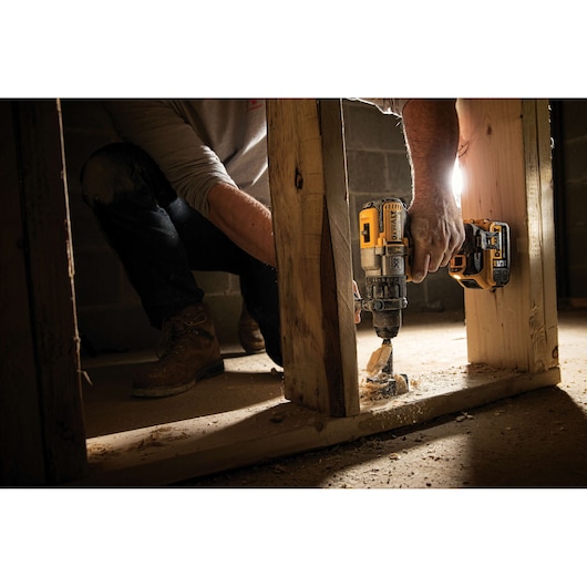 A person using a DEWALT DCD996P2 power drill to drill through a wooden beam at a construction site. There is sawdust on the floor and the person is wearing work boots and jeans.