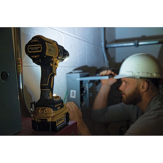 A close-up of a DeWalt cordless power drill resting on a surface in the foreground, with a person wearing a white hard hat working on an electrical box in the background.
