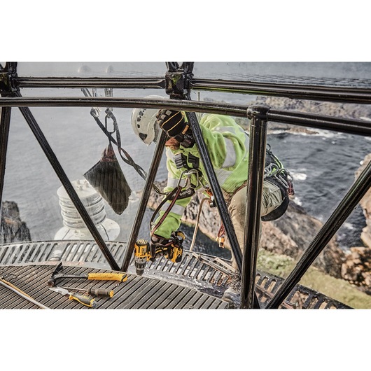 A worker wearing safety equipment and a helmet is using a DEWALT power drill on a metal structure near a rocky coastline. Several hand tools are placed on the metal platform, and the ocean is visible in the background.