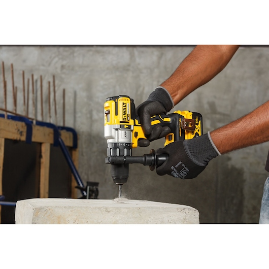 A person wearing black gloves uses a DEWALT cordless power drill to drill into a concrete block in an industrial construction setting.