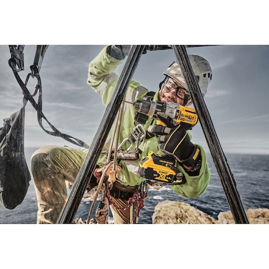 A worker equipped with safety gear uses a DEWALT DCD996P2 cordless power drill outdoors, positioned on a metal structure near a cliff with the sea in the background.