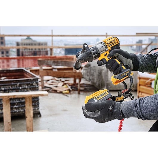 A person wearing gloves holds a DEWALT DCD996P2 cordless power drill with a battery pack, at a construction site with wooden structures and building materials in the background.