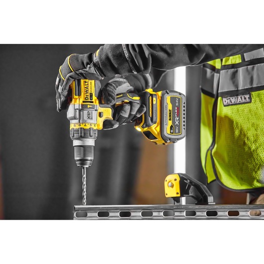 A person wearing gloves and a high visibility vest is using a DEWALT DCD999NT cordless drill to drill into a metal beam.