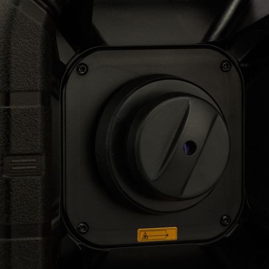 Close-up image of a black DEWALT device showing a circular knob or lens cover, mounted on a square panel with visible screws and a yellow caution sticker below.