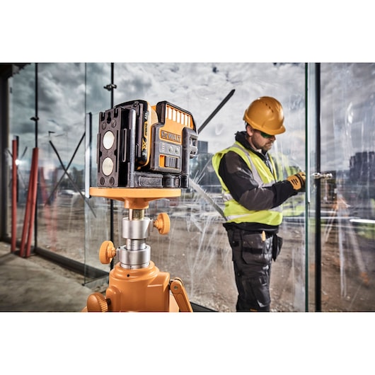 A DEWALT DCE0825D1G laser level mounted on a tripod is positioned in the foreground at a construction site, with a worker wearing a yellow hard hat and high-visibility vest using tools near glass windows in the background.