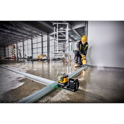 A DEWALT green line laser level tool (model DCE089D1G18) is set up on a construction site floor, projecting green laser lines to align metal tracks. A construction worker in protective gear and a yellow helmet is working with a power tool near a wall. Industrial scaffolding and various construction materials are visible in the background.