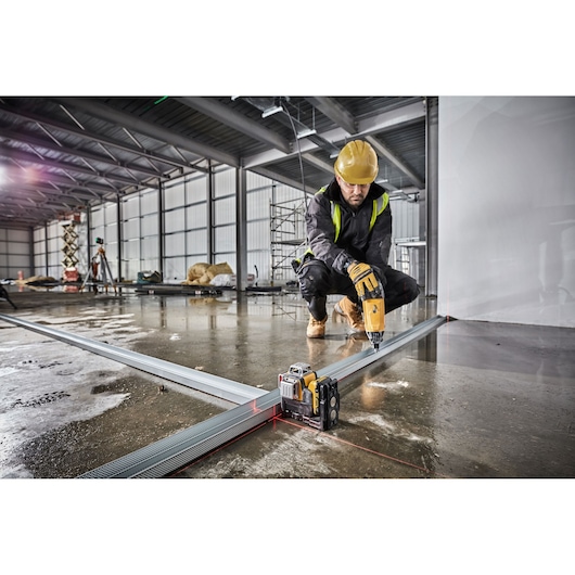 A construction worker using a Dewalt laser level (DCE089D1R) to align metal framing on a wet concrete floor in an industrial setting. The worker is wearing a yellow hard hat and operating a power drill while kneeling.