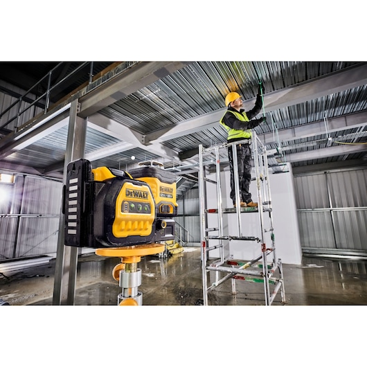 A DEWALT DCE089NG18 laser level is mounted on a tripod inside an industrial building under construction. In the background, a worker wearing a hard hat and safety vest stands on a scaffold, installing wiring along the metal ceiling beams.
