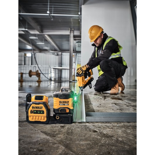 A DEWALT 12V/18V green cross line laser level is shown on a construction site floor alongside another DEWALT measuring tool. In the background, a construction worker wearing personal protective equipment including a hard hat, gloves, and high-visibility vest, is kneeling and using a DEWALT power tool on a metal frame.