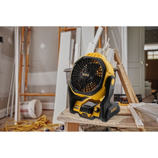 A DEWALT portable battery-powered fan placed on a wooden workbench in a construction or renovation environment, surrounded by tools, wooden planks, and construction materials.
