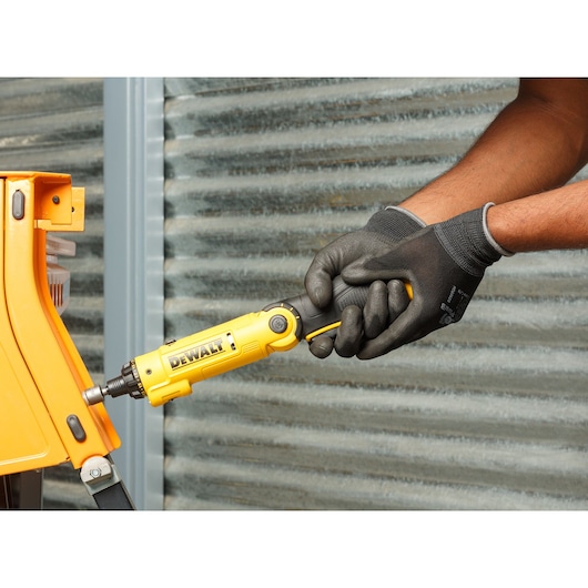 A person wearing black protective gloves is using a yellow DEWALT cordless screwdriver (model DCF008) to fasten a screw on an orange piece of equipment. Corrugated metal wall is visible in the background.